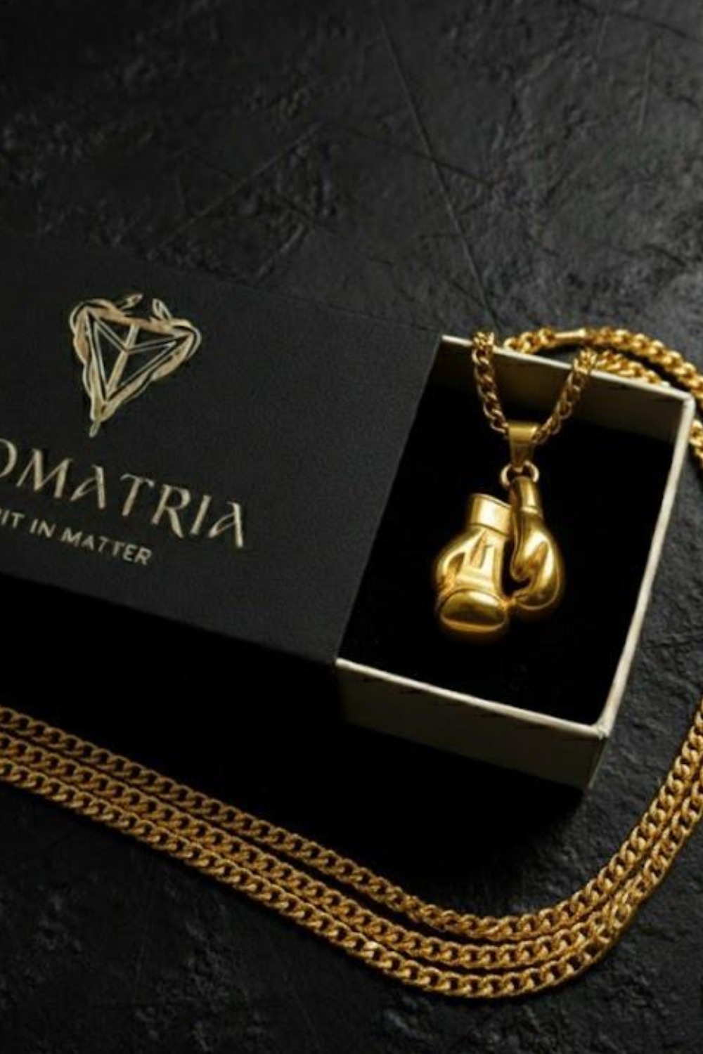 A gold chain necklace with a pendant in the shape of boxing gloves, placed inside a black box with a gold chain.