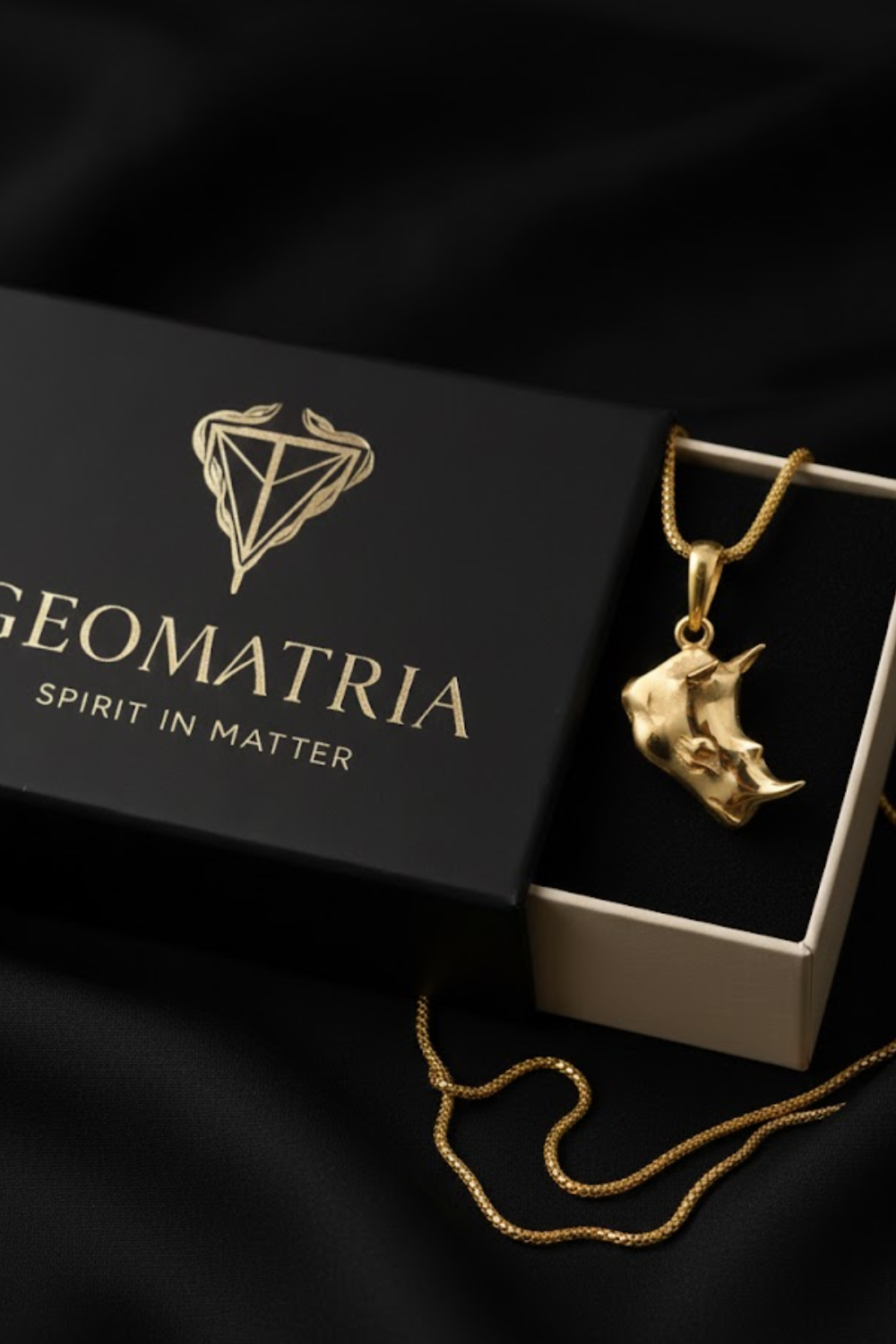 A gold necklace with a pendant is displayed in a black box with the text "Geomatria" and "Spirit in Matter" printed on it.