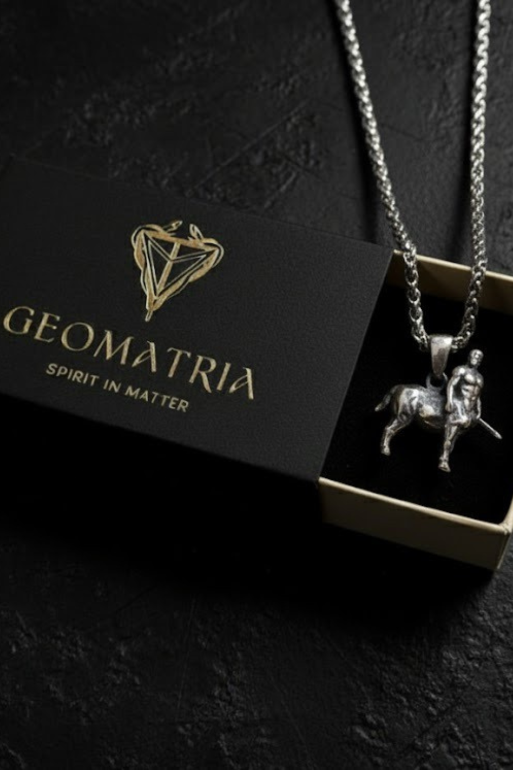 A silver necklace with a pendant is displayed in a black box with the text "Geomatria" and "SPIRIT IN MATTER" printed on it.