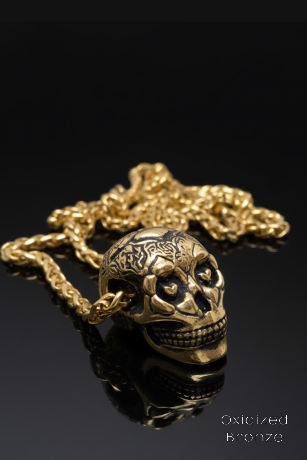 a gold-colored pendant in the shape of a skull, with intricate designs and patterns, resting on a reflective surface.