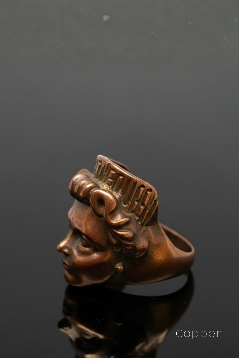A copper-colored ring with a profile view of a woman's face, featuring a crown-like design on the top.