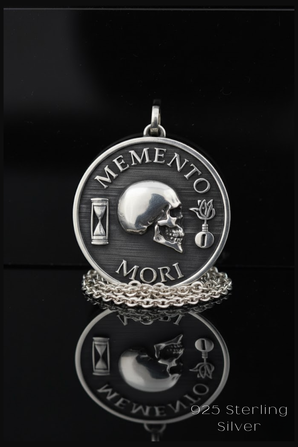A silver-colored pendant with the text "MEMENTO MORI" and a skull design, along with a small flower and a sand timer, is displayed against a black background.