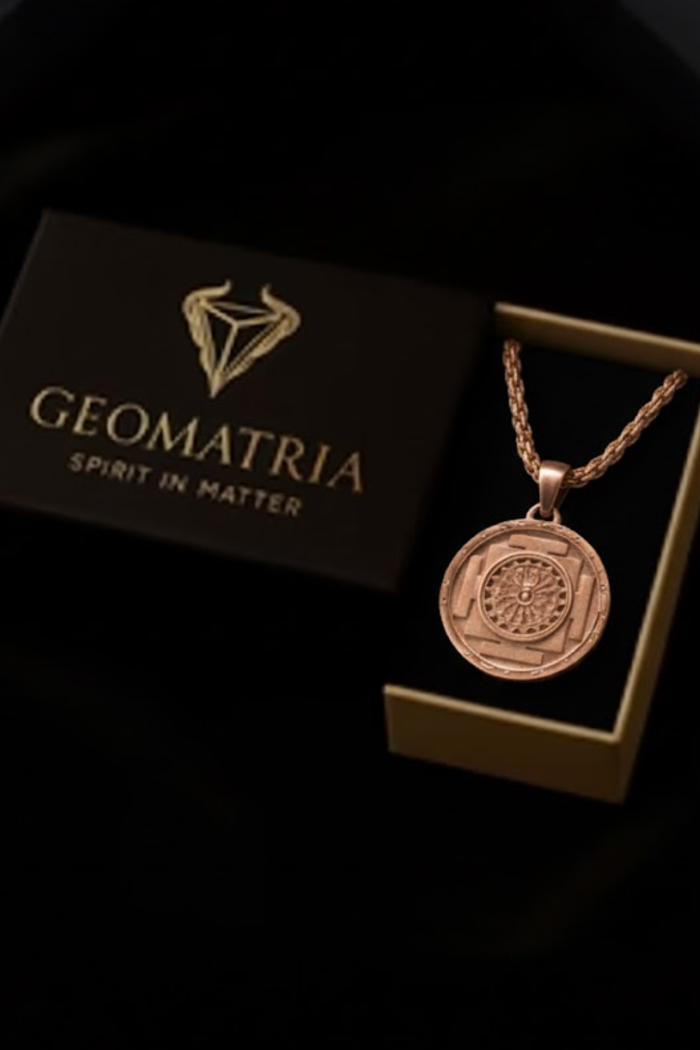 A gold-colored pendant necklace with a circular medallion-like design is displayed in a black box with the text "Geomatria" and "Spirit in Matter" printed on it.