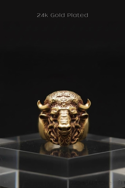 A 24k gold-plated ring featuring a detailed buffalo head design is displayed on a clear glass stand against a black background.