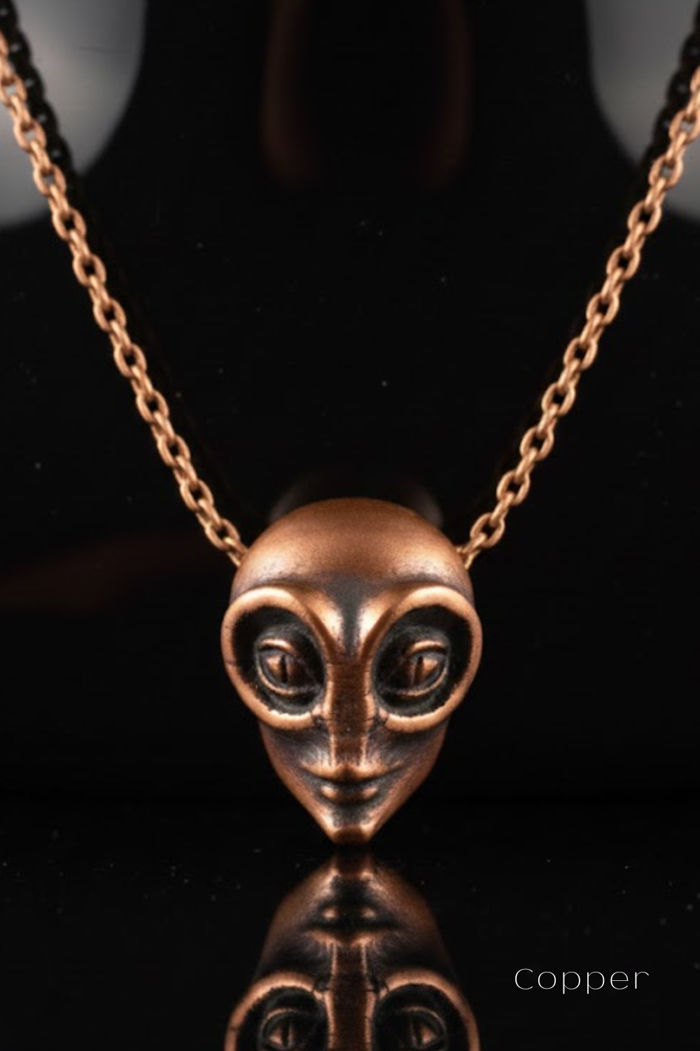 A copper-colored pendant necklace with a unique alien-like face design, displayed against a black background.