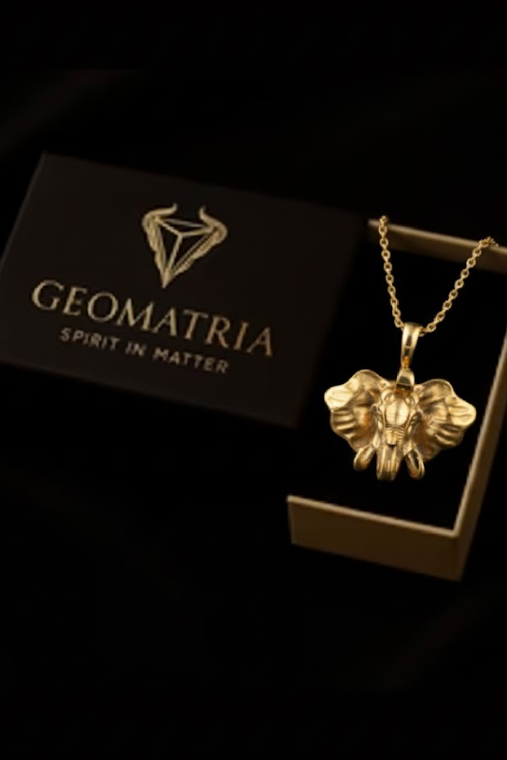 A gold elephant pendant necklace is displayed in a black box with the text "Geomatria Spirit in Matter" printed on it.