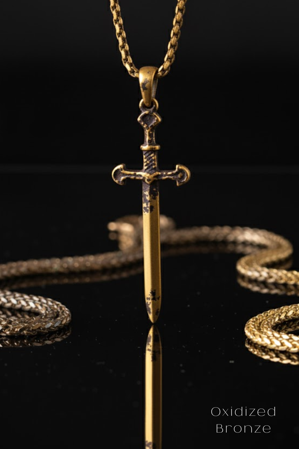 A close-up view of a gold-colored sword pendant hanging from a chain against a black background.