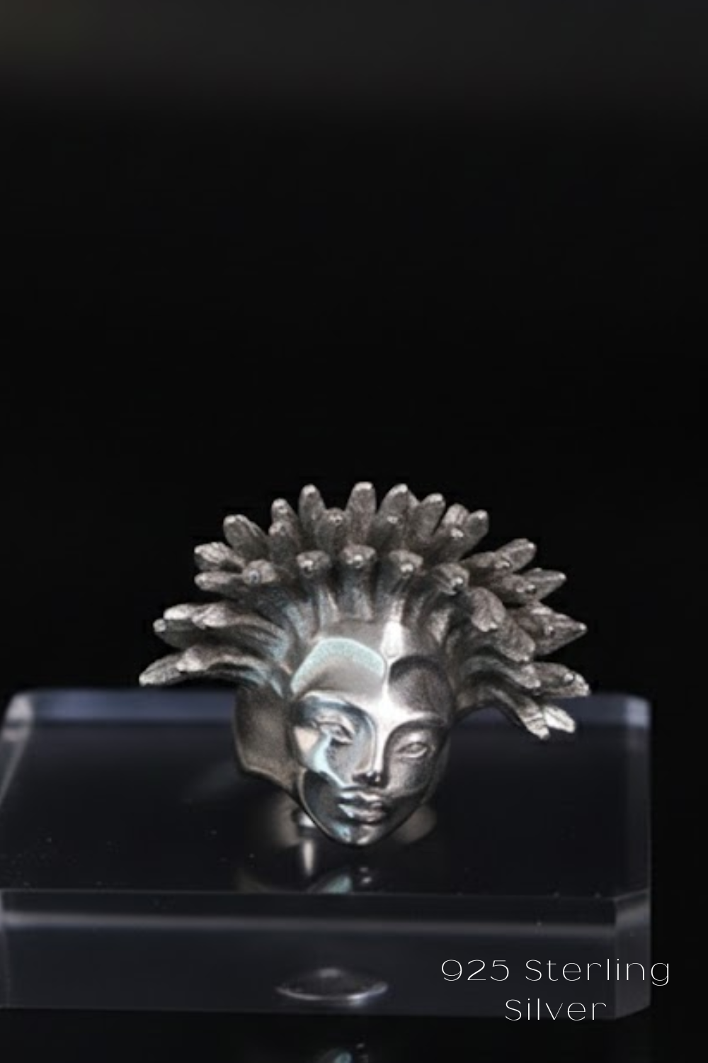 A silver-colored metal sculpture of a face with a large, intricate hairstyle is displayed on a clear, transparent stand against a black background.