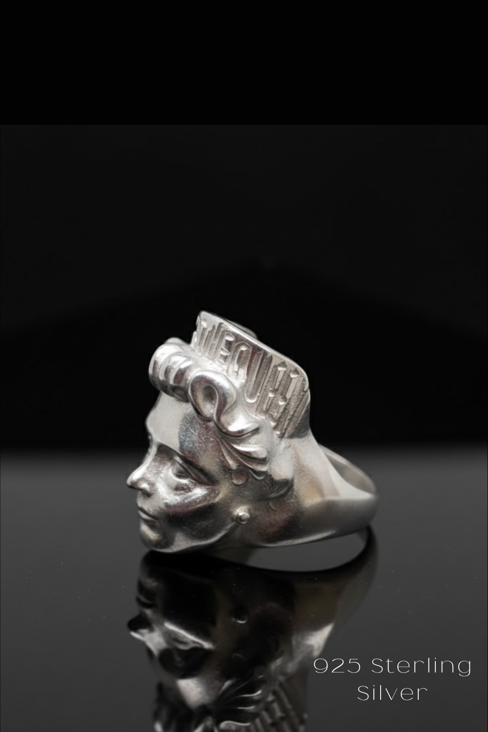 A silver ring with a face design on it, placed on a reflective surface against a black background.