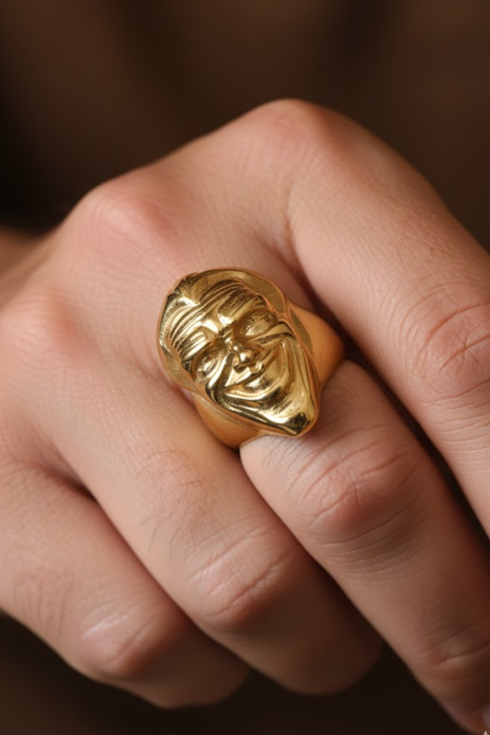 A close-up image of a person's hand wearing a gold ring with a face design on it.