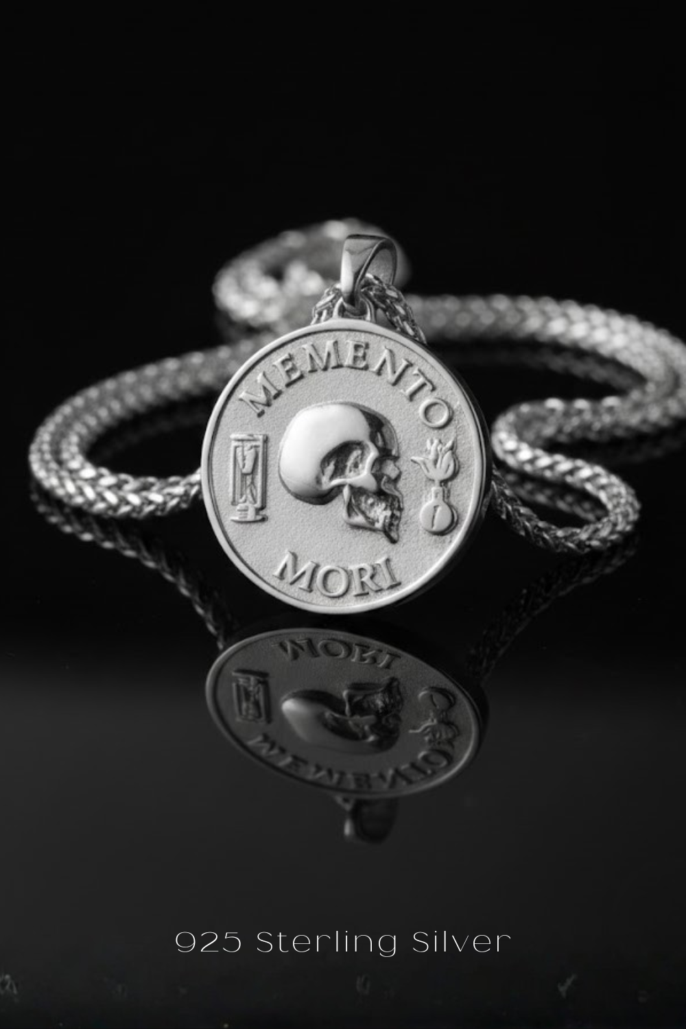 A silver medallion pendant with the Latin motto "Memento mori" engraved on it, hanging from a chain against a black background.