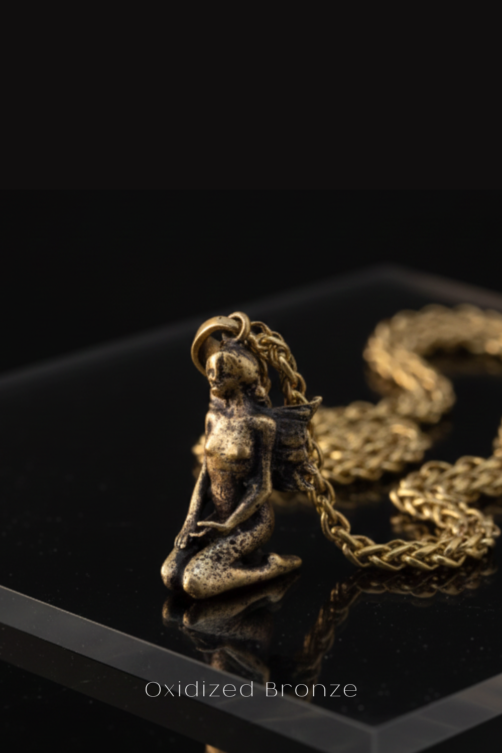 A close-up image of a small, bronze-colored pendant or charm in the shape of a woman, sitting on a reflective surface.