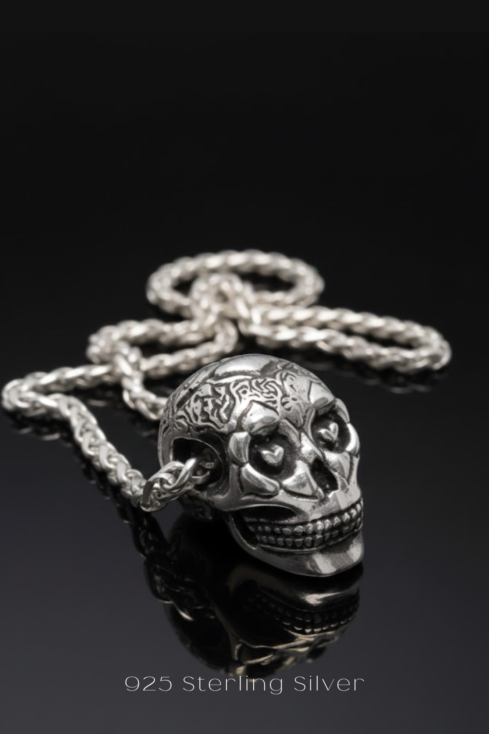 A silver-colored pendant in the shape of a skull, with intricate designs and a chain attached to it, resting on a reflective surface.