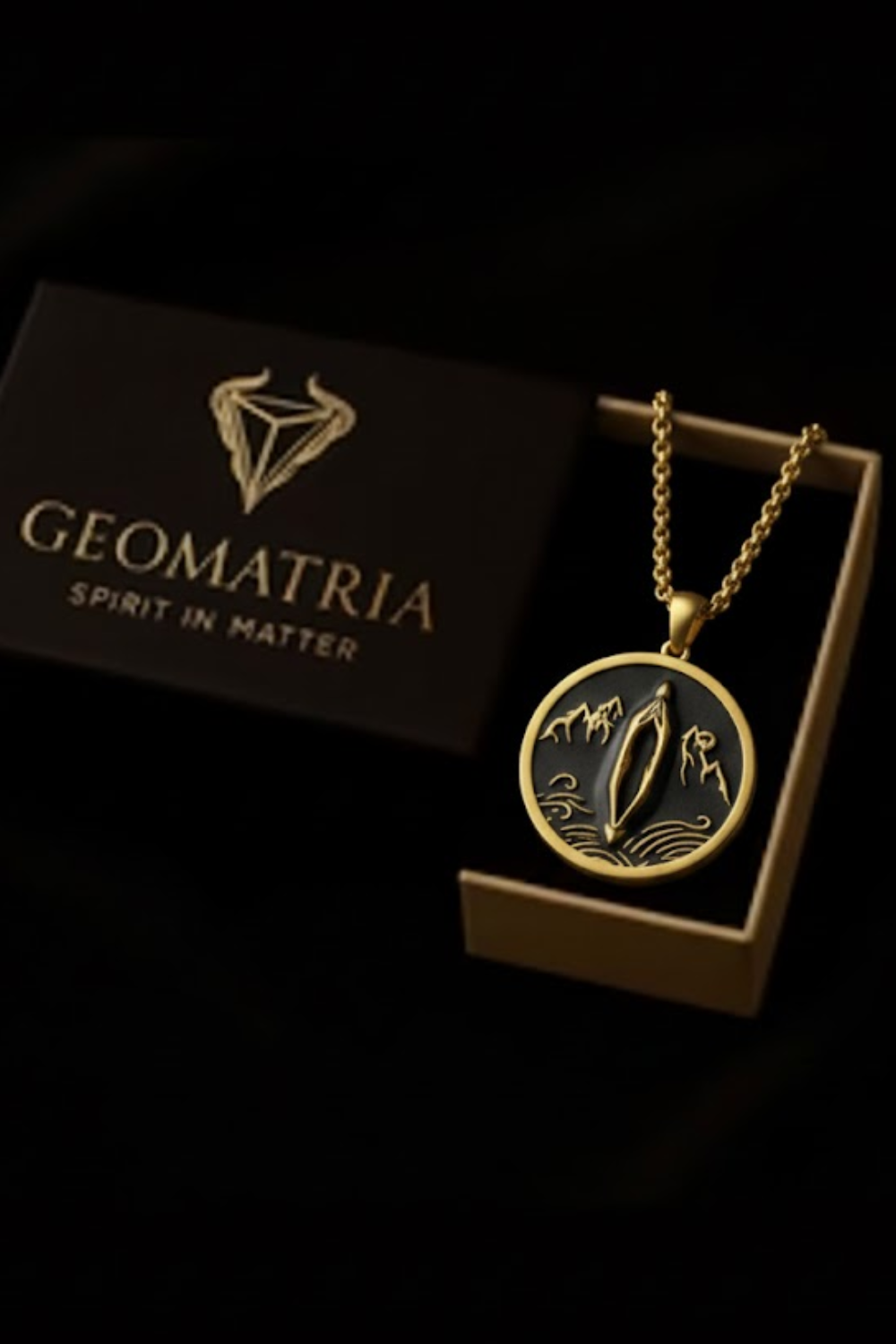 A gold necklace with a pendant featuring a design of mountains and a river, displayed in a black box with the text "Geomatria" and "Spirit in Matter" printed on it.