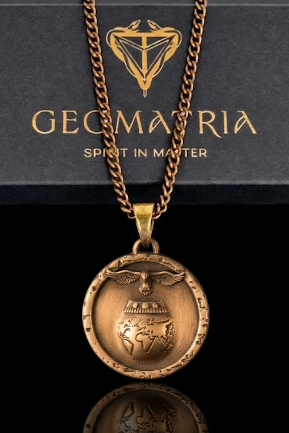 A gold-colored pendant necklace with a circular medallion featuring a bird and a globe design, hanging against a black background with a gold-colored logo.