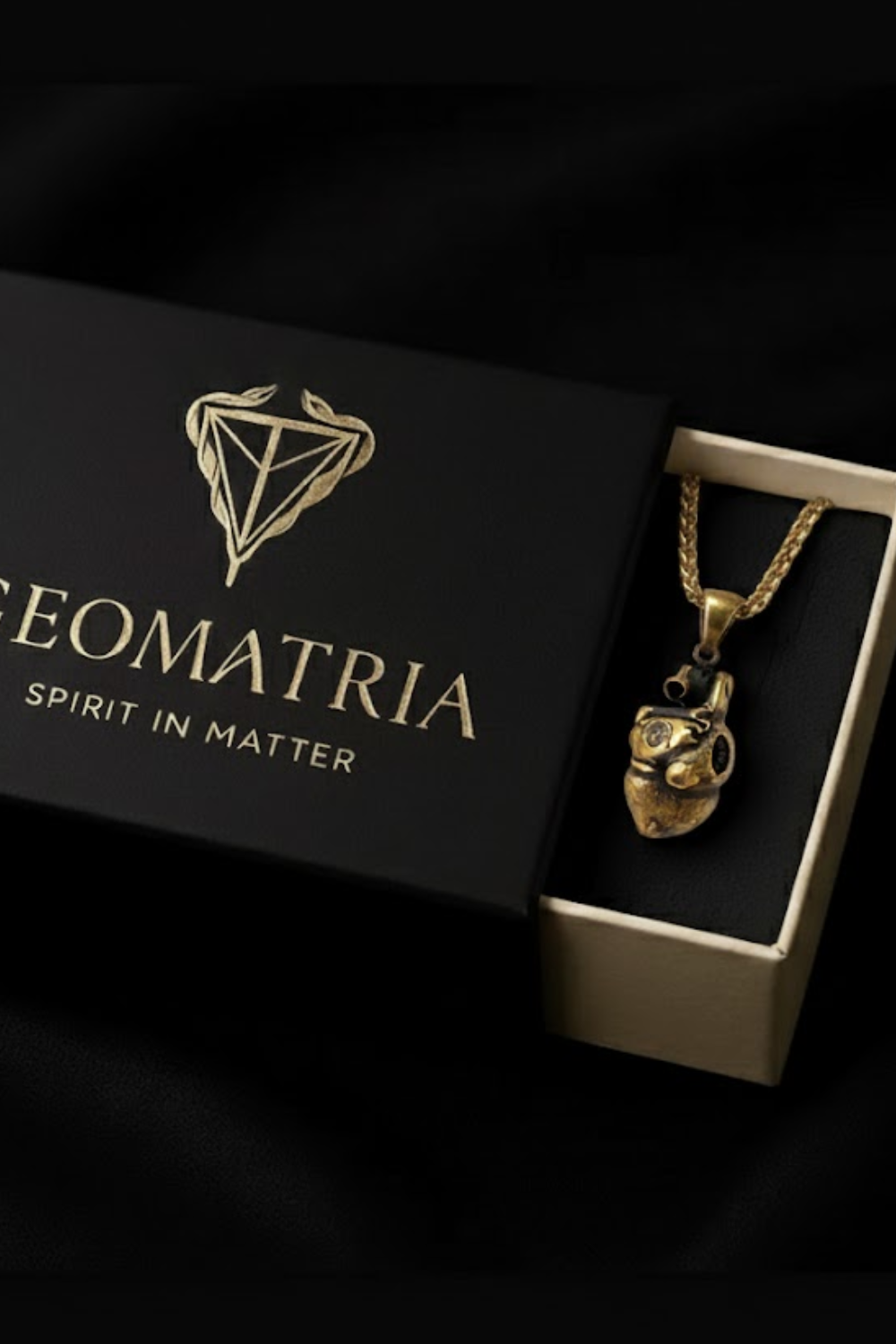 A gold-colored necklace with a pendant is displayed in a black box with the text "Geomatria" and "SPIRIT IN MATTERS" printed on it.