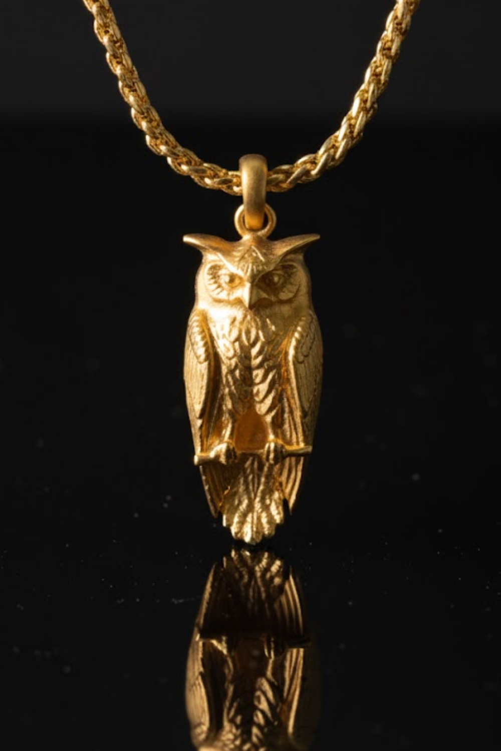 A gold-colored pendant in the shape of an owl is displayed against a black background, with its reflection visible below it.