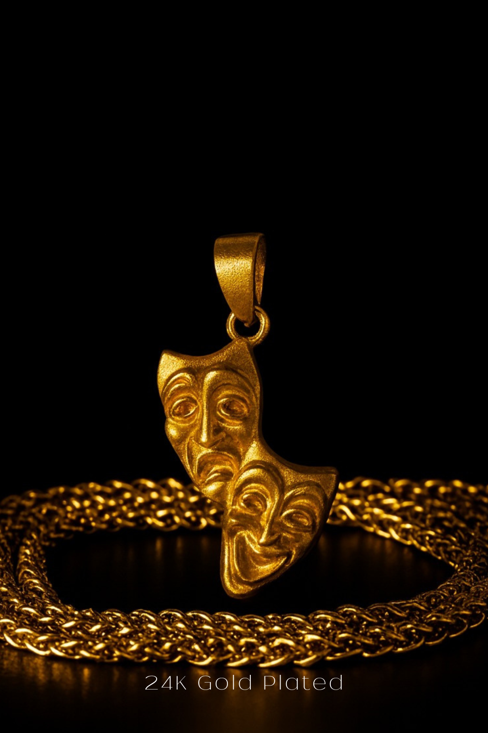 A 24k gold-plated pendant featuring two golden theatrical masks is displayed against a black background, with a golden chain necklace visible in the foreground.