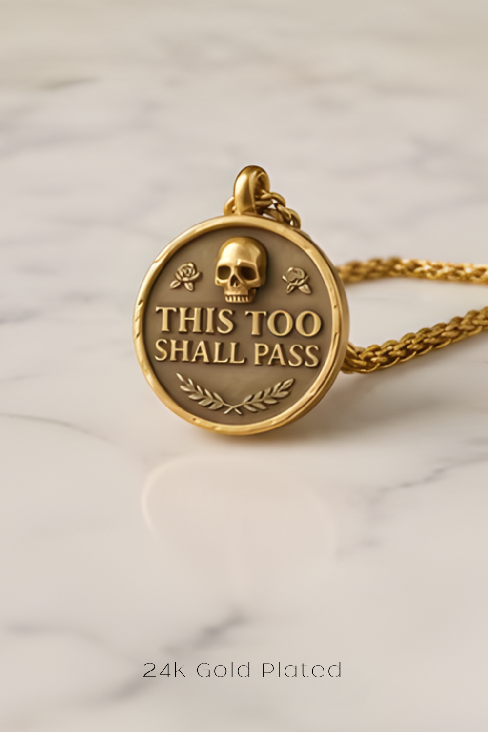 A gold-plated pendant with the text "THIS TOO SHALL PASS" and a skull design, resting on a marble surface.