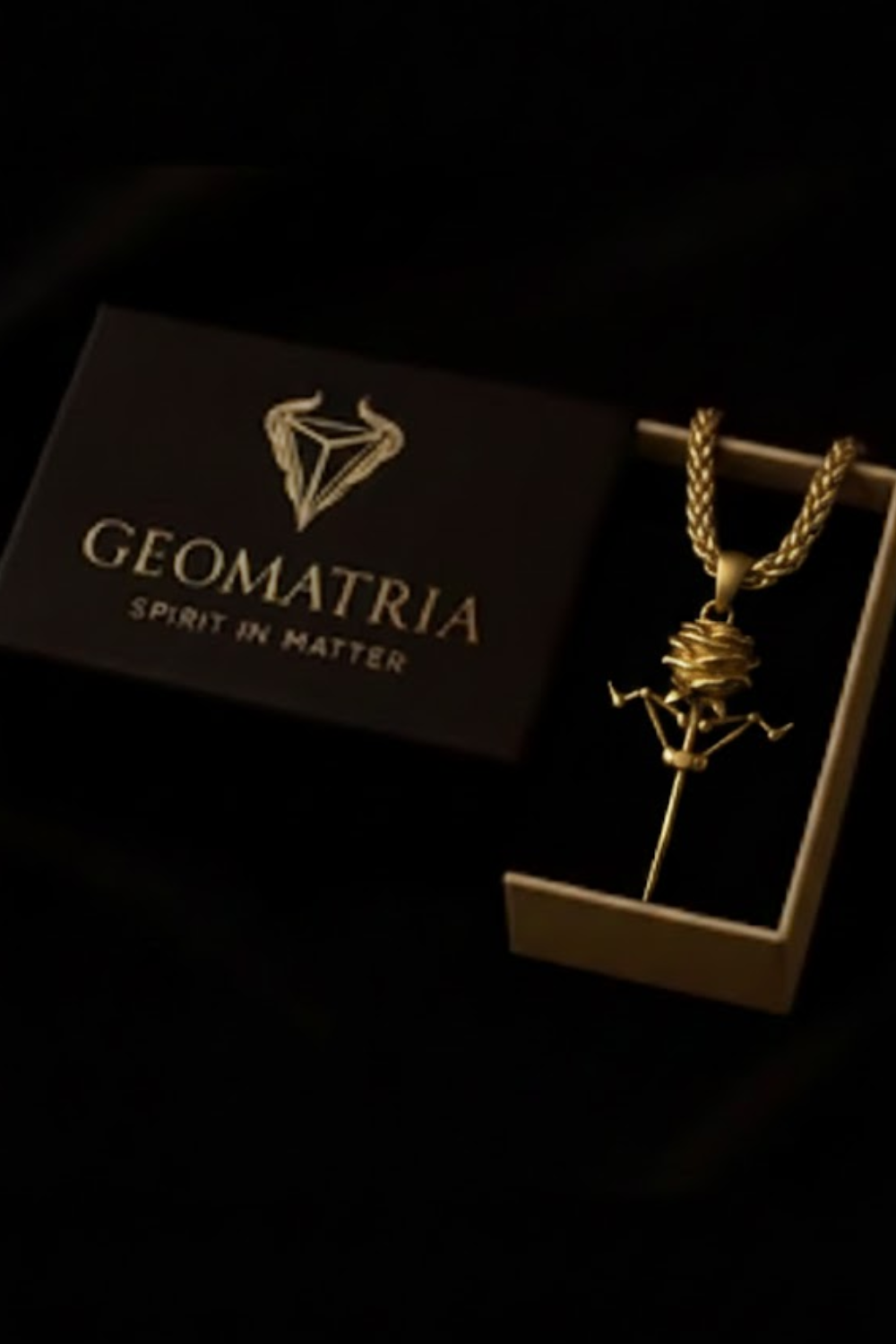 A gold necklace with a pendant is displayed in a black box with the text "Geomatria" and "Spirit in Matter" printed on it.