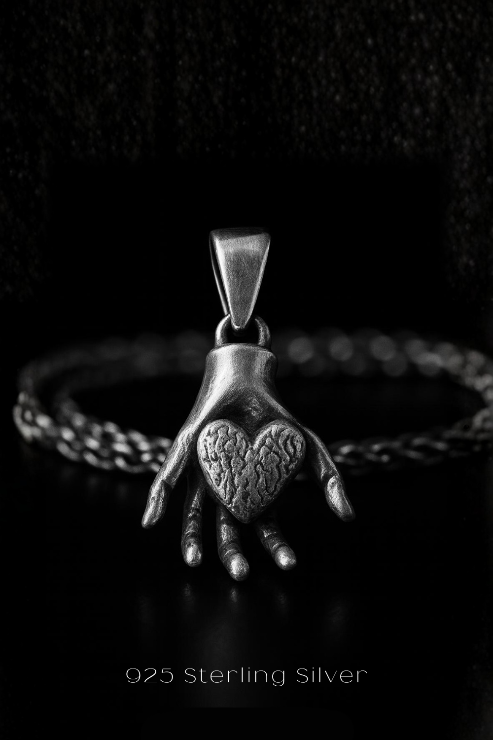 A silver pendant in the shape of a hand holding a heart, with a chain around it, against a black background.