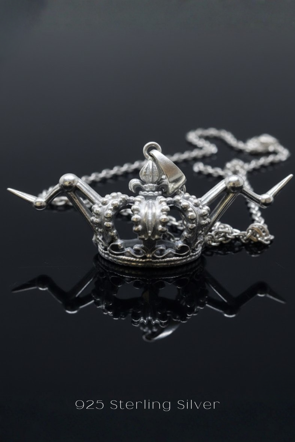 A silver-colored pendant necklace with a crown-shaped pendant is displayed against a dark background.