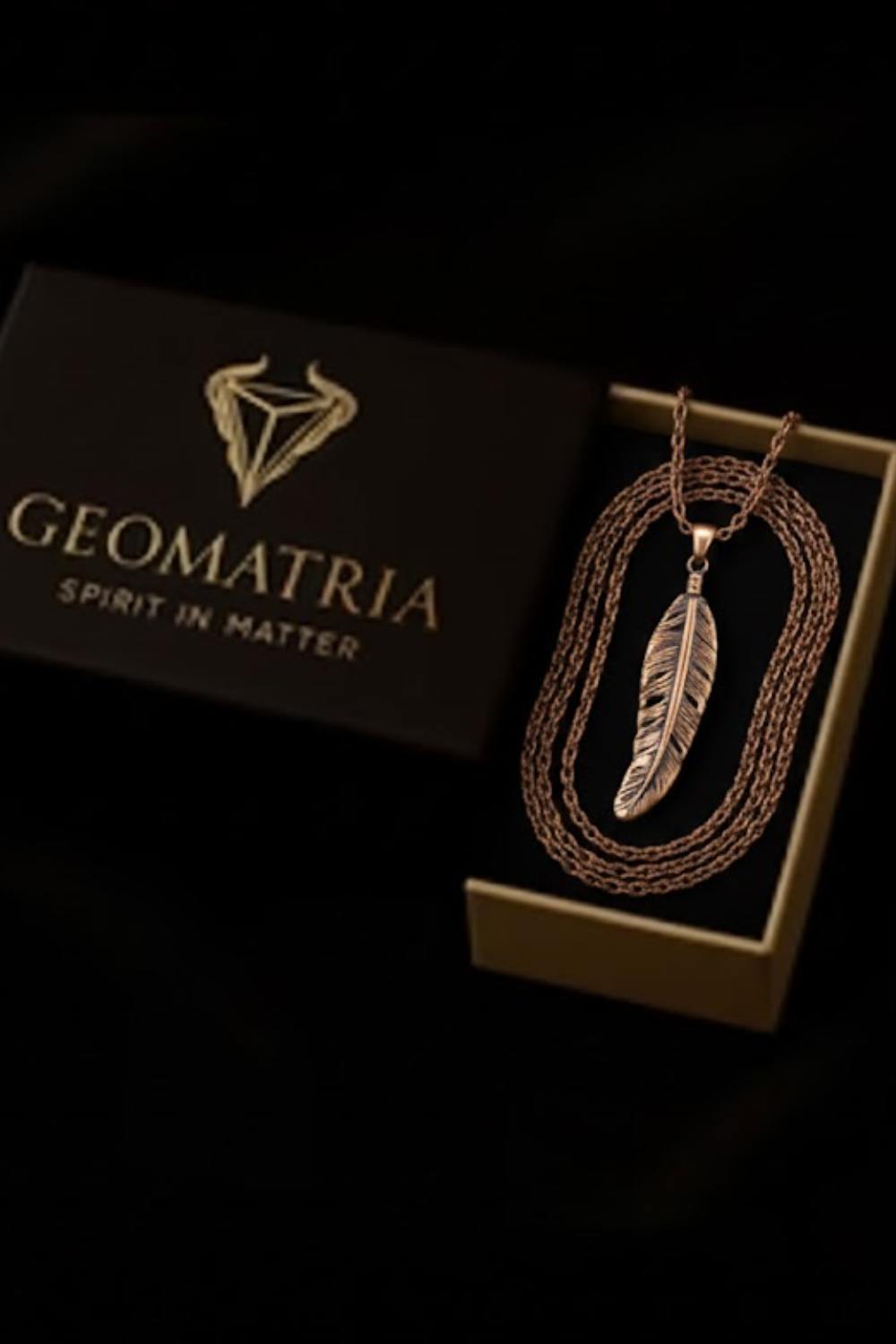 A gold-colored feather pendant necklace is displayed in a black box with the text "Geomatria Spirit in Matter" printed on it.
