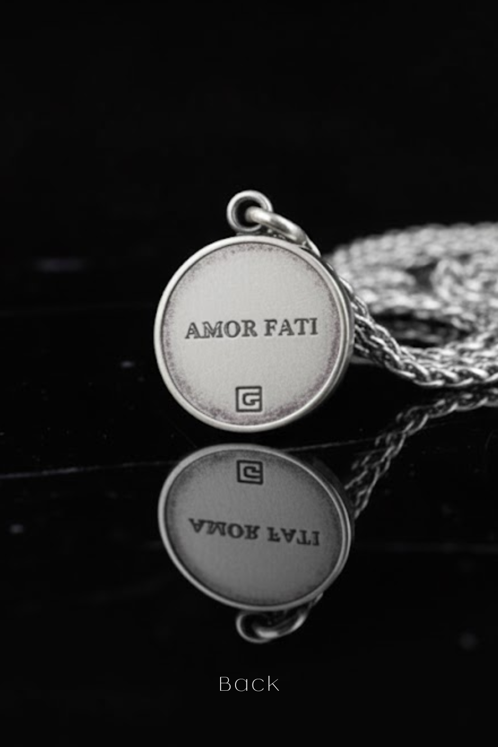 a silver pendant with the text "Amor Fati" engraved on it, resting on a reflective surface.