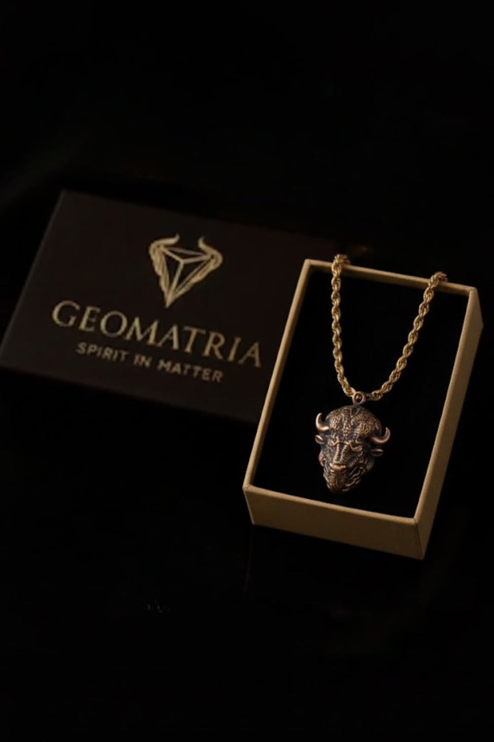 A gold-colored necklace with a pendant in the shape of a bull's head is displayed in a box with the Geomatria logo.