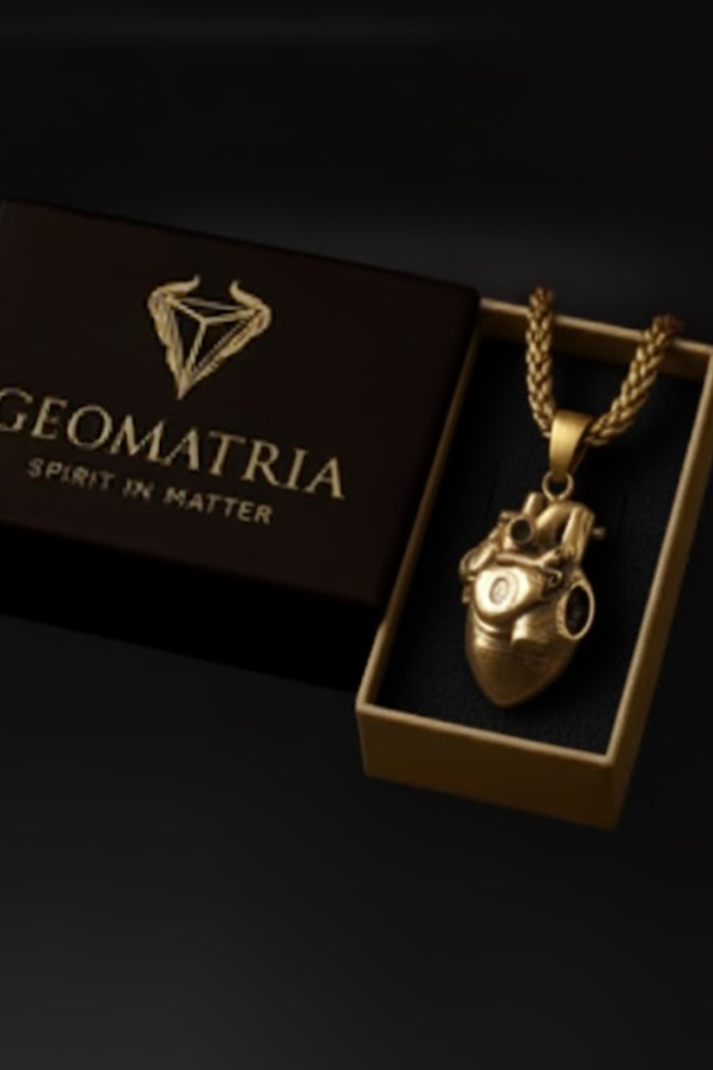 A gold-colored heart-shaped pendant necklace is displayed in a black box with the text "Geomatria" and "SPIRIT IN MATTERS" printed on it.