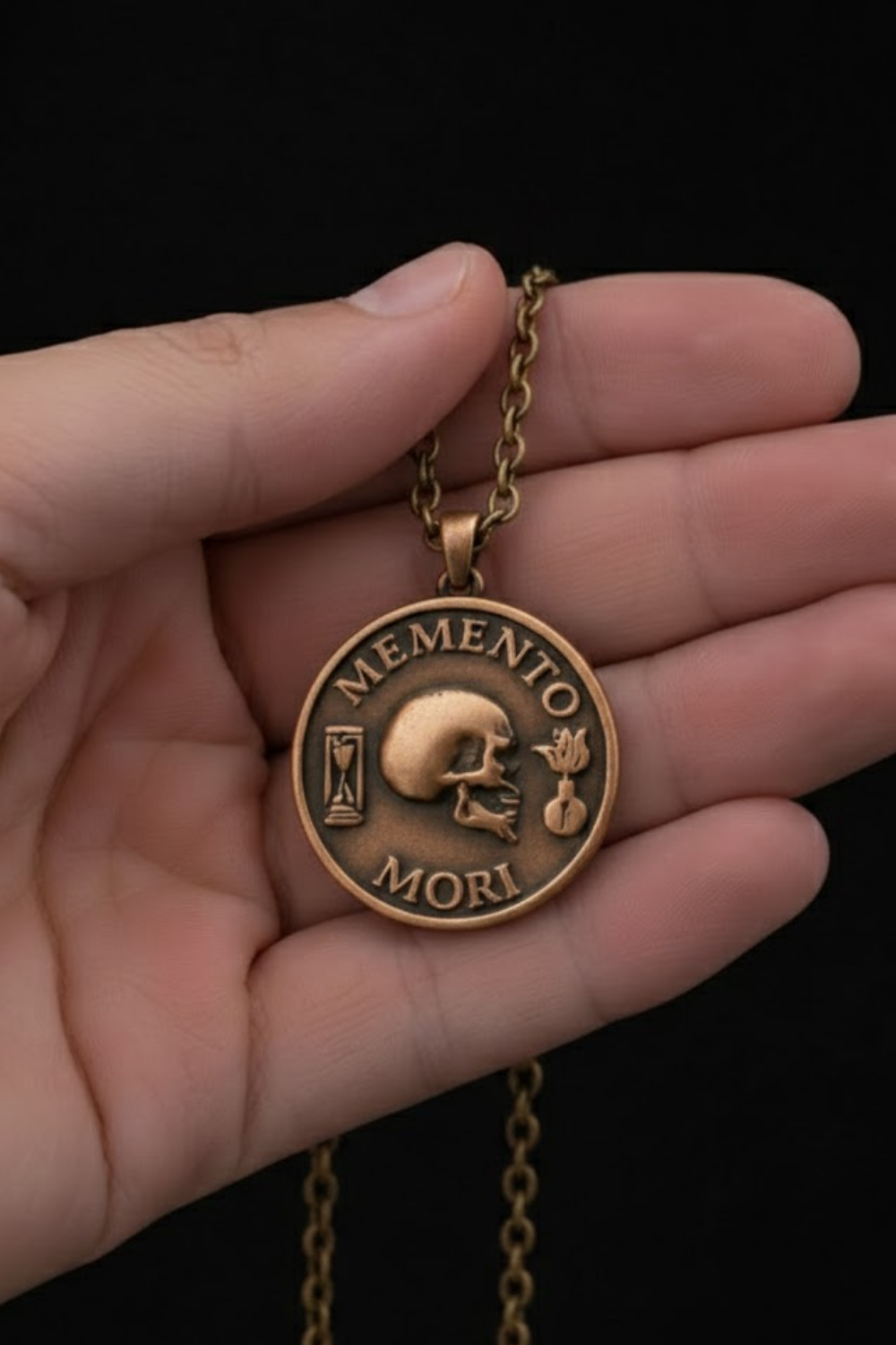 A hand holding a gold-colored pendant necklace with a circular medallion featuring a skull and the Latin phrase "Memento mori" (which means " remembrance of death ").