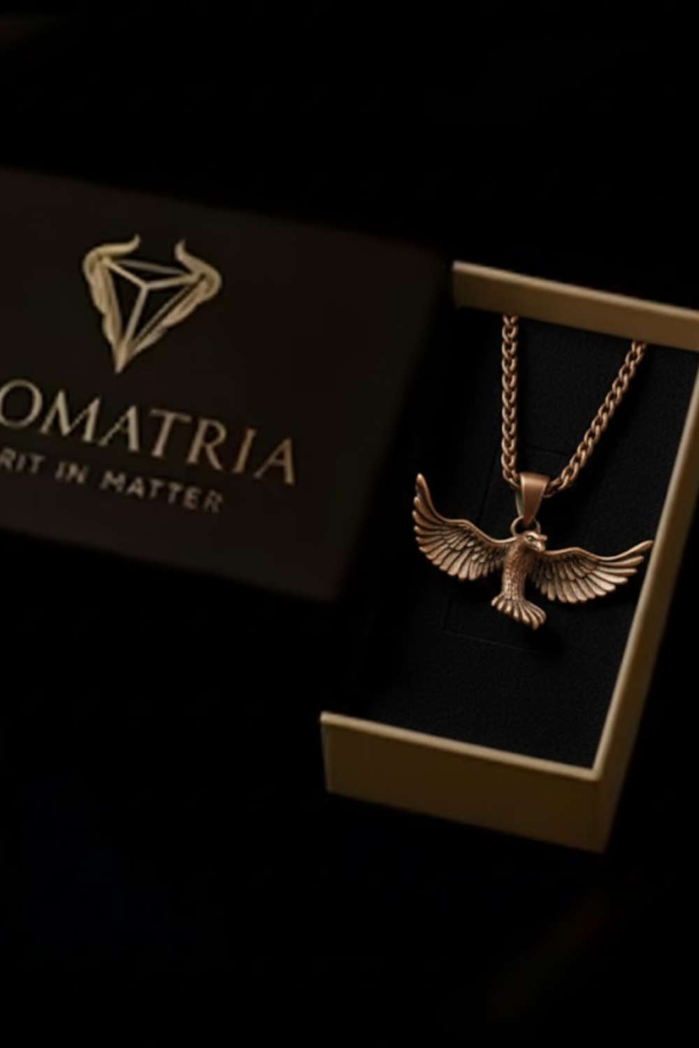 A gold-colored pendant necklace with a winged design is displayed in a black box with the word "OMATRIIA" written on it.