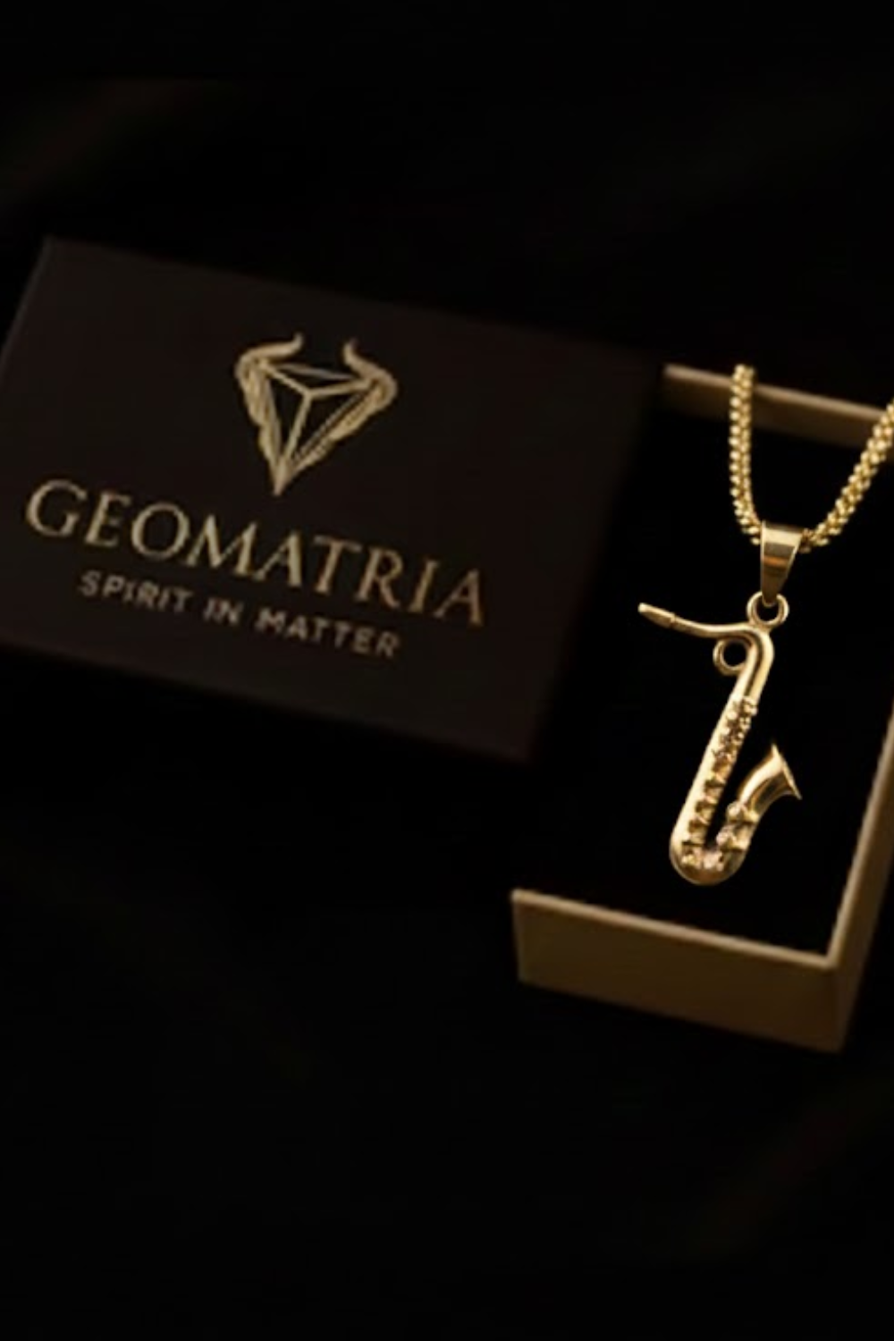 A gold saxophone pendant is displayed next to a black box with the word "Geomatria" printed on it.