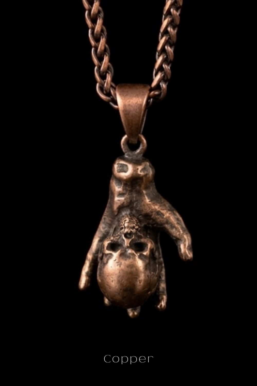 A copper-colored pendant necklace with a unique, sculpted design featuring a hand-like figure holding a skull-like object.