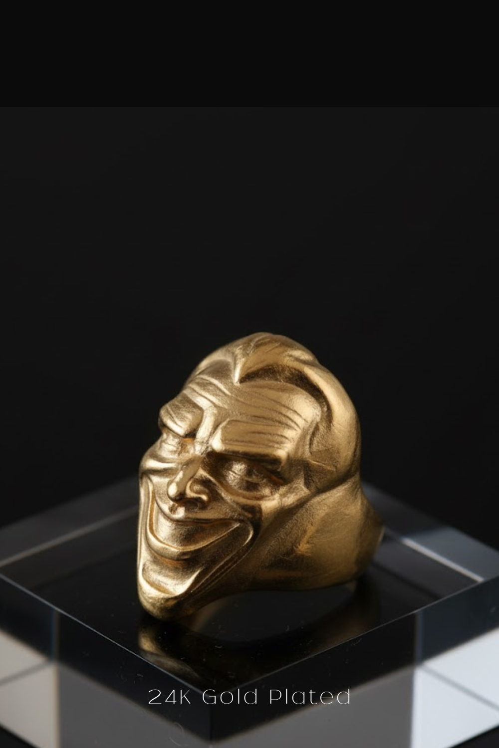 A 24K gold-plated ring in the shape of a smiling, exaggerated face is displayed against a black background.