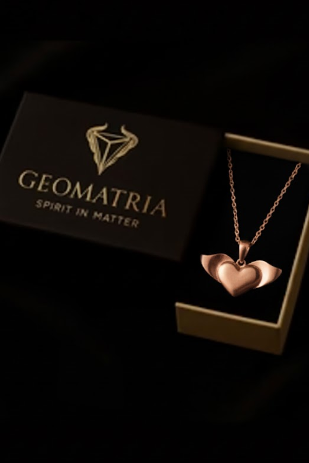 A necklace with a heart-shaped pendant is displayed in a gold box with the text "Geomatria" and "SPIRIT IN MATERIA" printed on it.