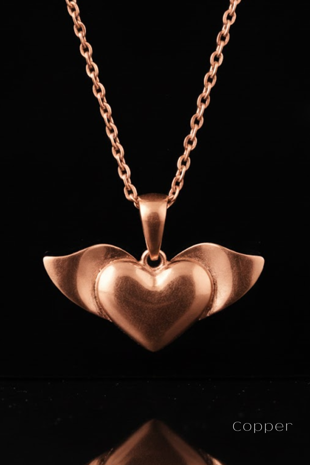 A copper-colored heart-shaped pendant with wings attached, suspended from a chain against a black background.