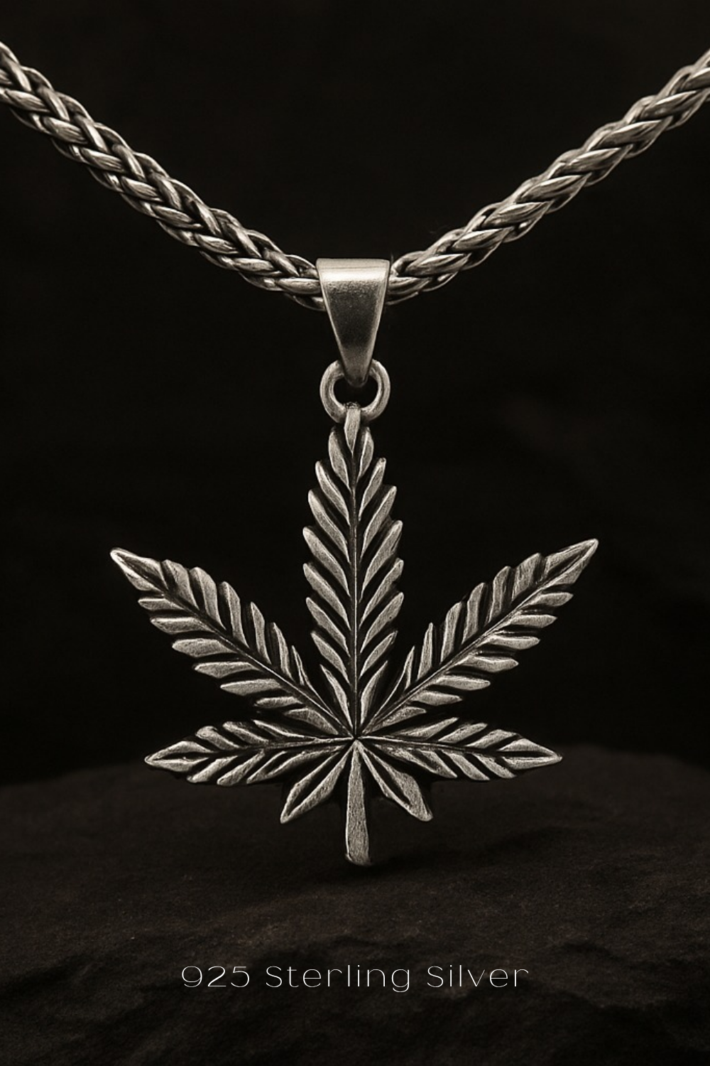 A silver pendant in the shape of a leaf, possibly a marijuana leaf, is suspended from a chain against a dark background.