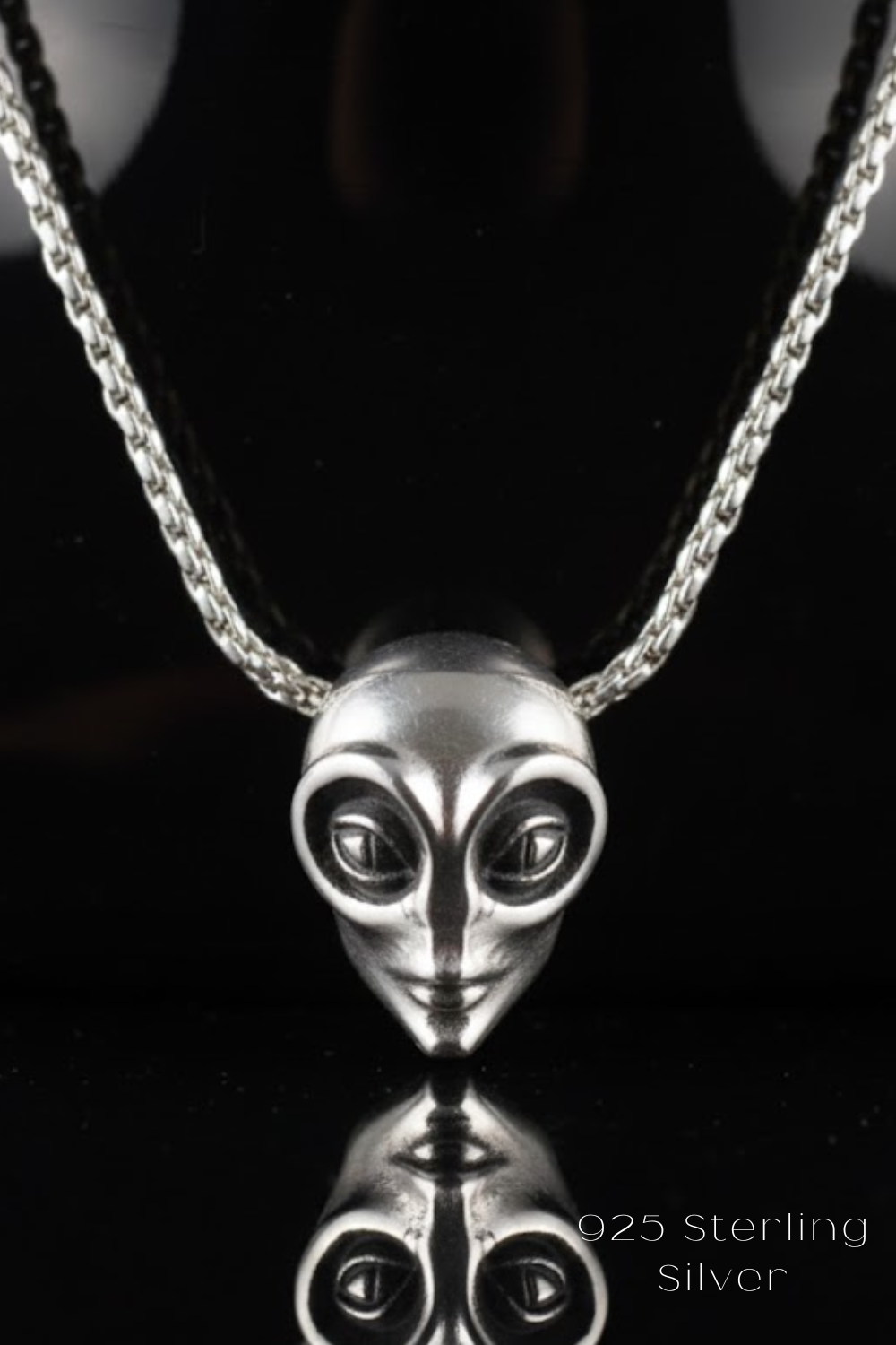 A silver necklace with a pendant in the shape of an alien face, reflecting on a black surface.