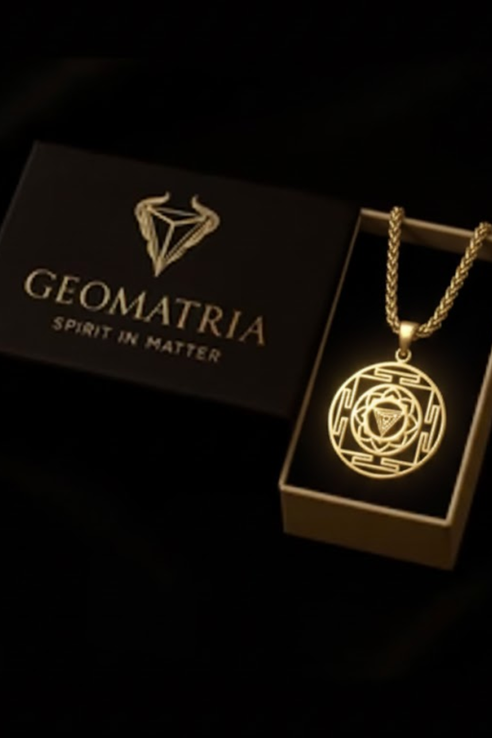 A gold necklace with a pendant is displayed in a black box with the Geomatria logo, accompanied by a gold chain.