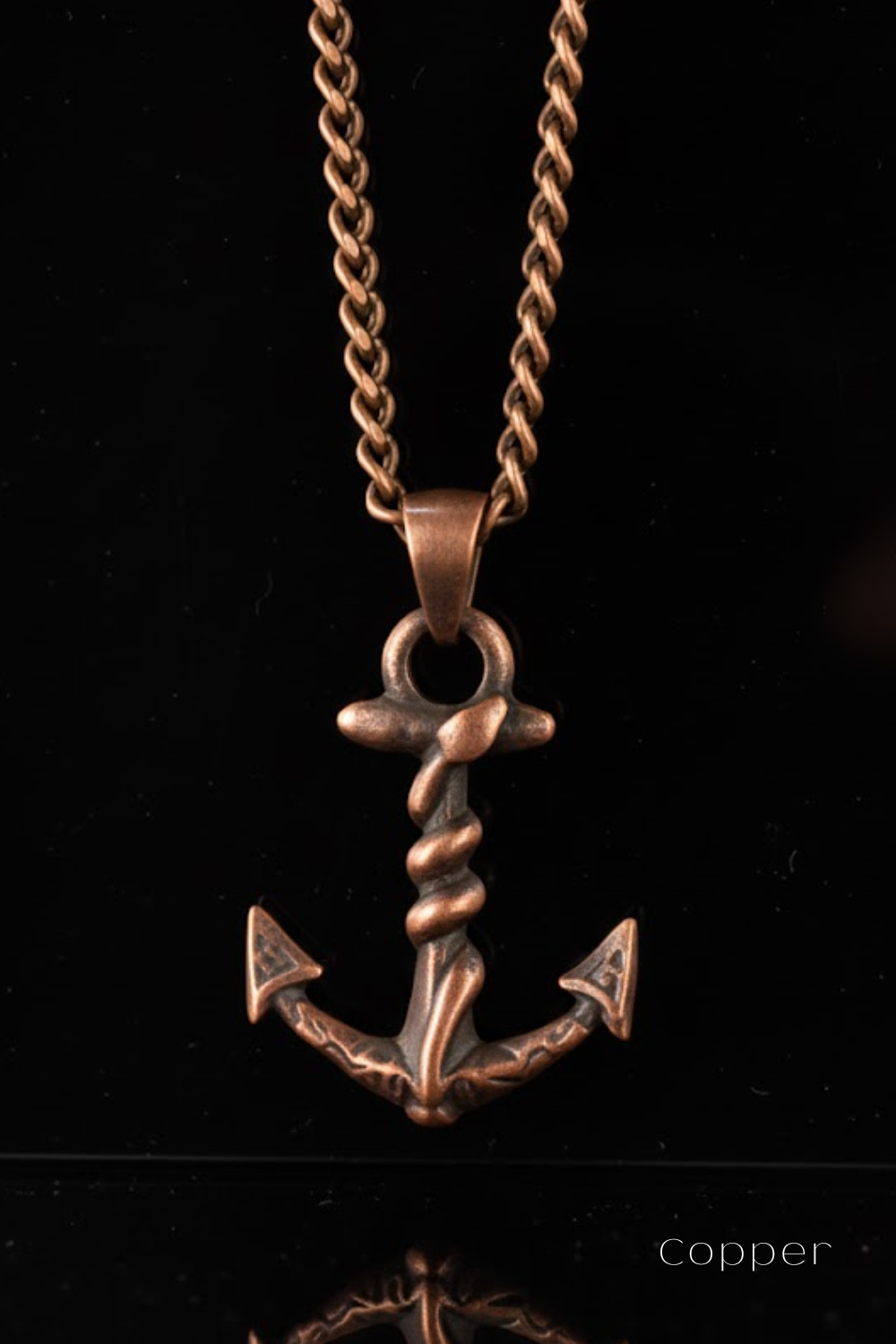 A copper-colored metal anchor pendant with a twisted design, suspended from a chain against a black background.