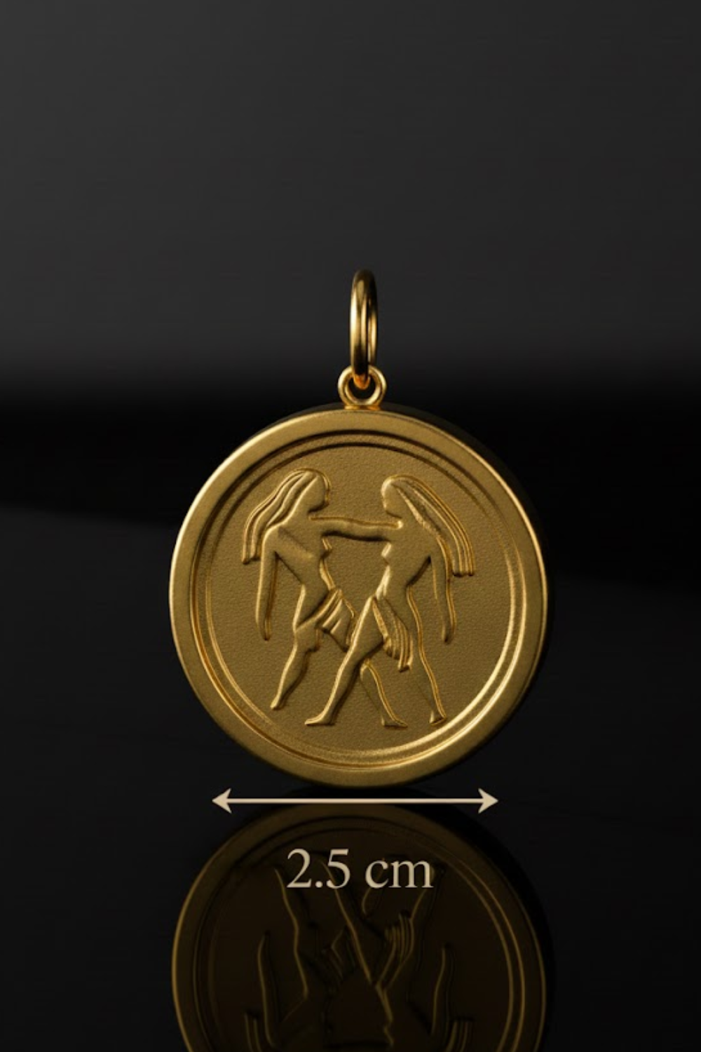 A gold-colored pendant with an embossed design of two figures, possibly representing the zodiac signs Aries and Taurus. The pendant is placed on a reflective surface, and there is a measurement scale indicating its size, which is 2.5 cm.