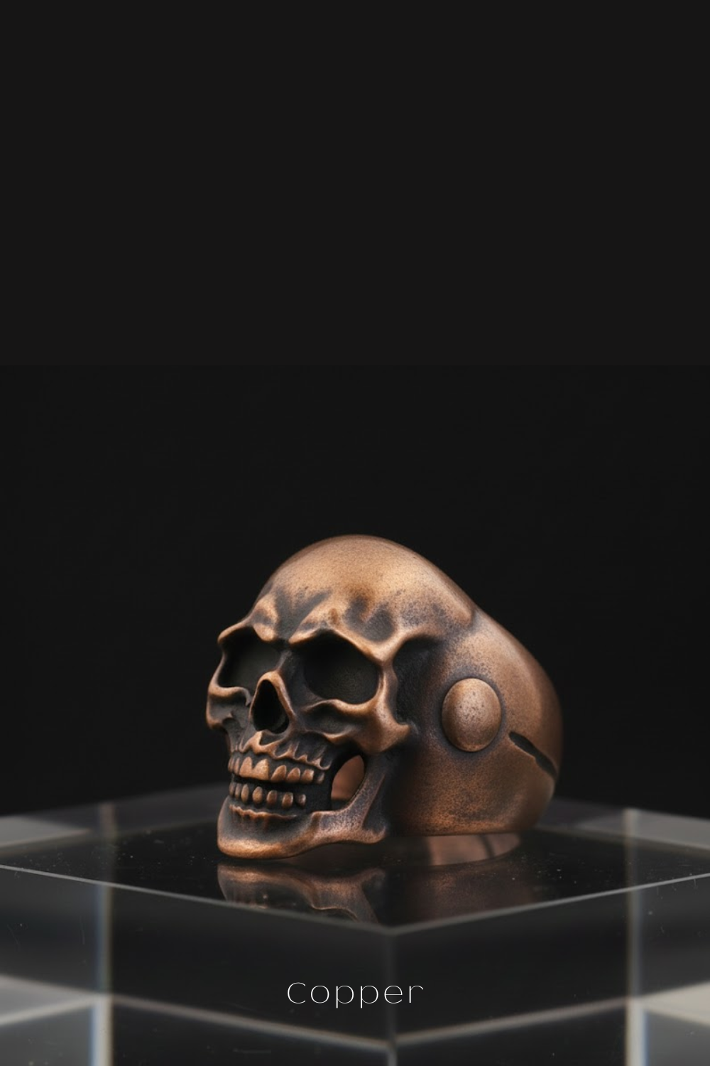A copper-colored metal skull ring is displayed on a clear glass stand against a black background.