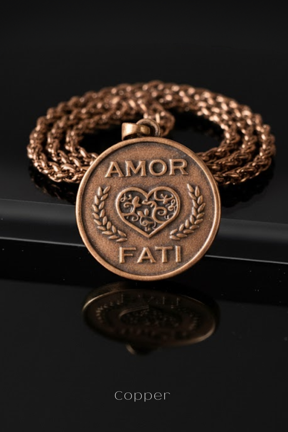 a close-up of a copper-colored pendant with the words "Amor Fati" engraved on it, which means "love is a feast" in Italian. The pendant is displayed on a chain against a black background.