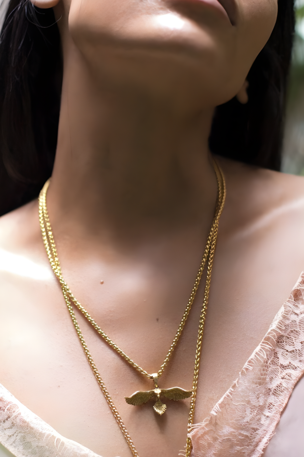 A close-up of a woman's neck and chest area, with a gold chain necklace and a pendant featuring a bird design.