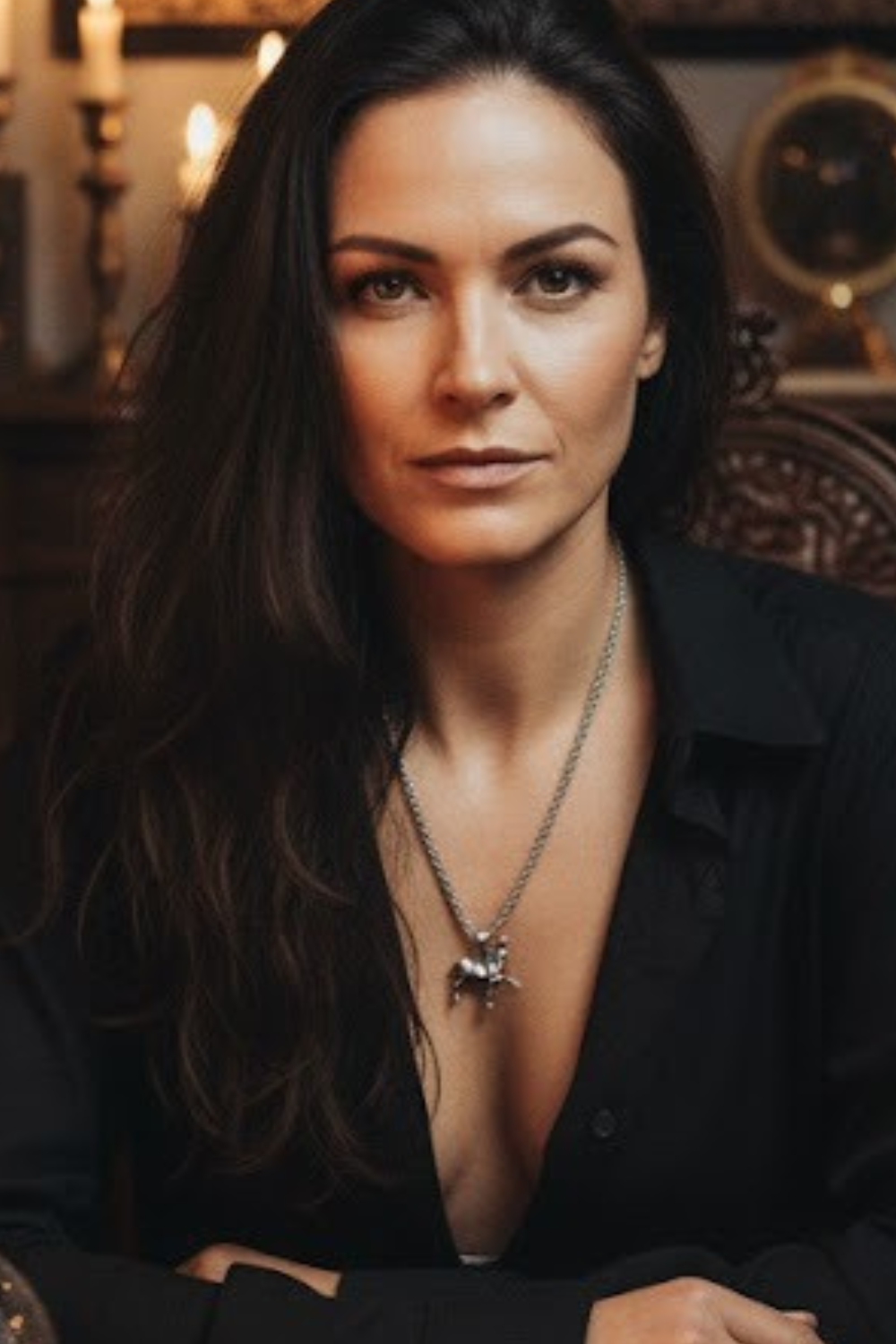 A woman with long dark hair wearing a black blazer and a necklace is sitting in a room with a clock and other objects in the background.