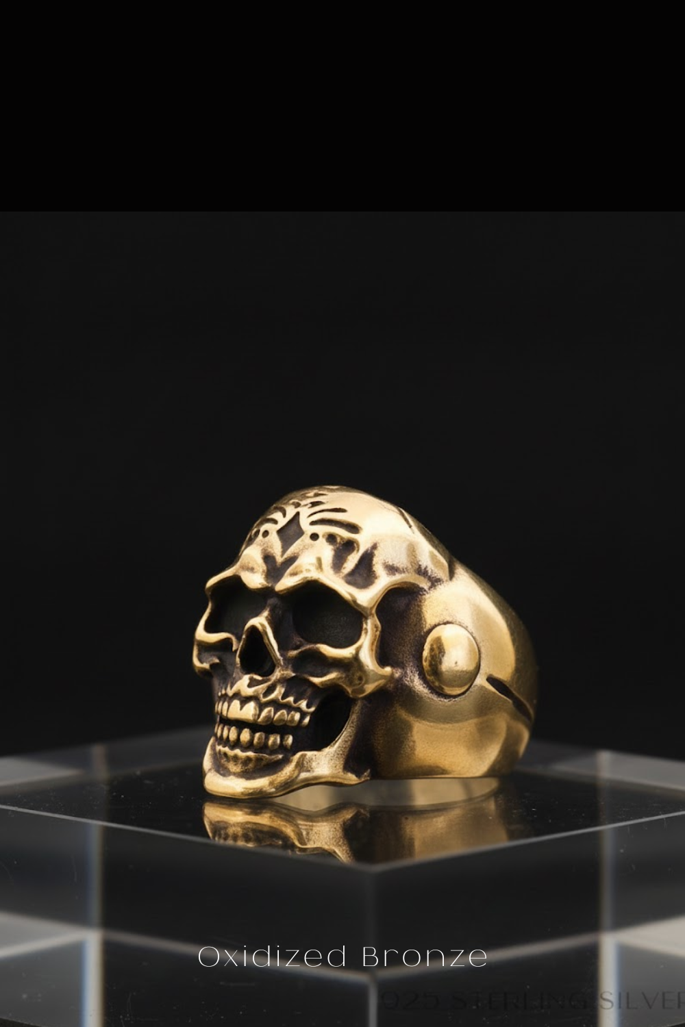 a gold-colored skull ring displayed on a clear glass stand against a black background.