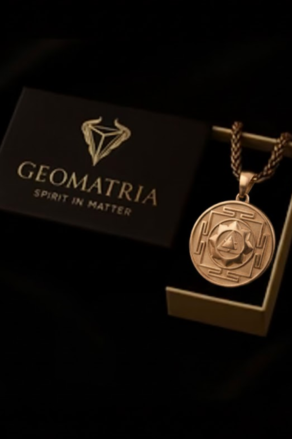 a gold-colored pendant with a geometric design, displayed in a black box with the text "Geomatria" and "SPIRIT IN MATTERS" printed on it.