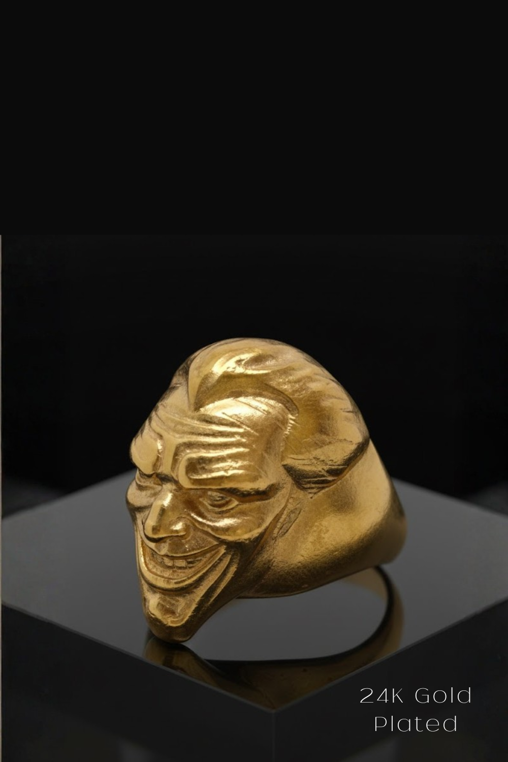 A 24K gold plated ring in the shape of a smiling man's face is displayed on a black pedestal against a black background.