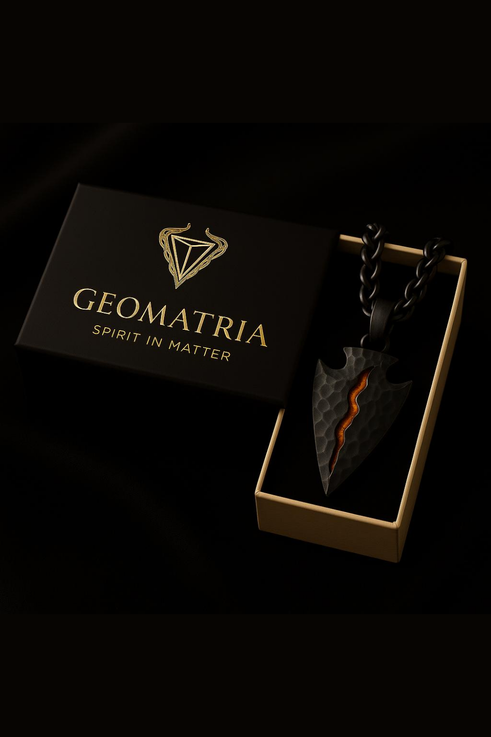 a black box with a gold logo and the text "Geomatria" on it, along with the phrase "SPIRIT IN MATTER". Next to the box is a necklace with a pendant in the shape of a flame or fire, which is placed inside a gold-colored box.